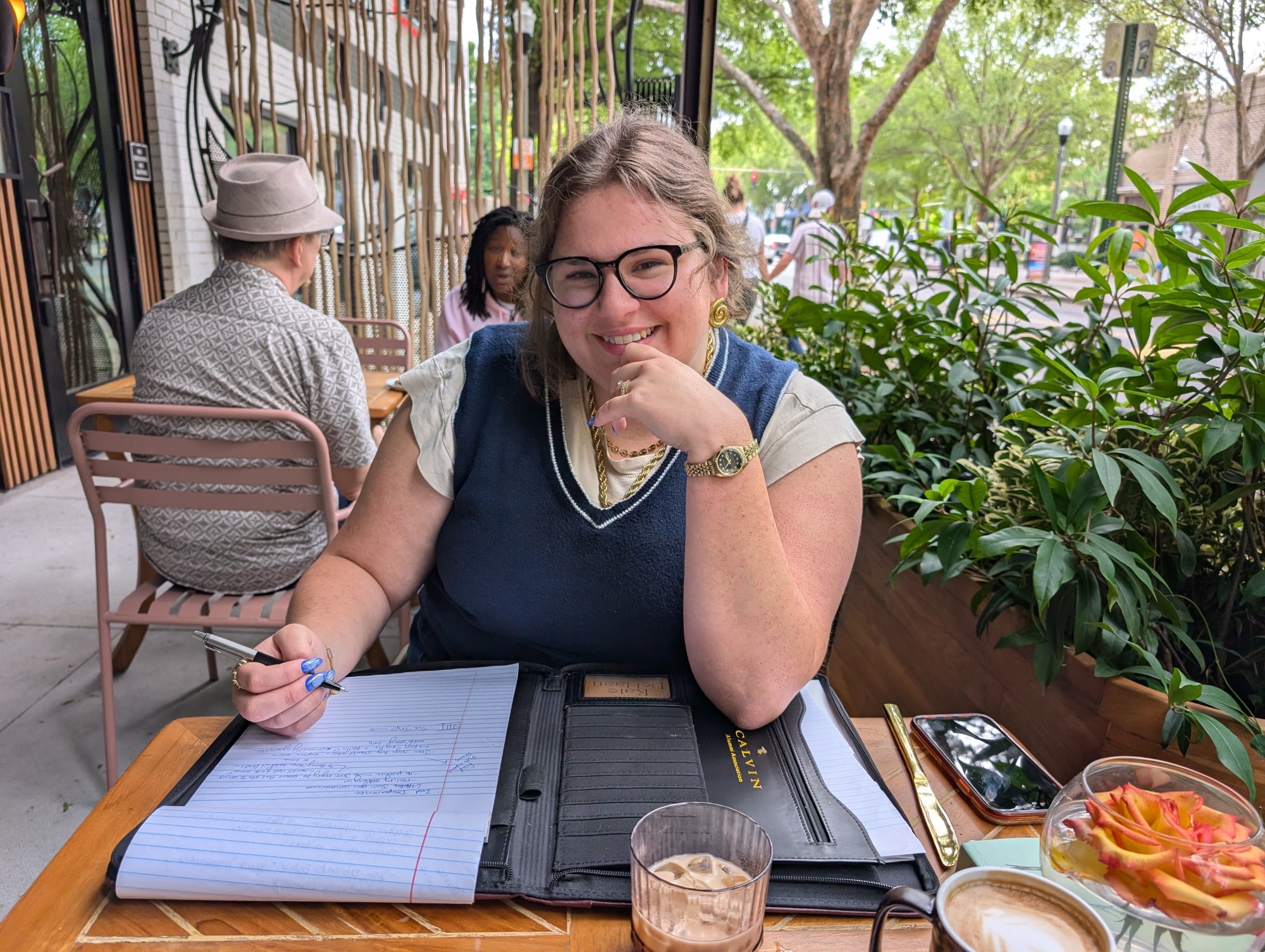 A smiling Kate is writing notes for her book in a legal pad at a coffee shop. 