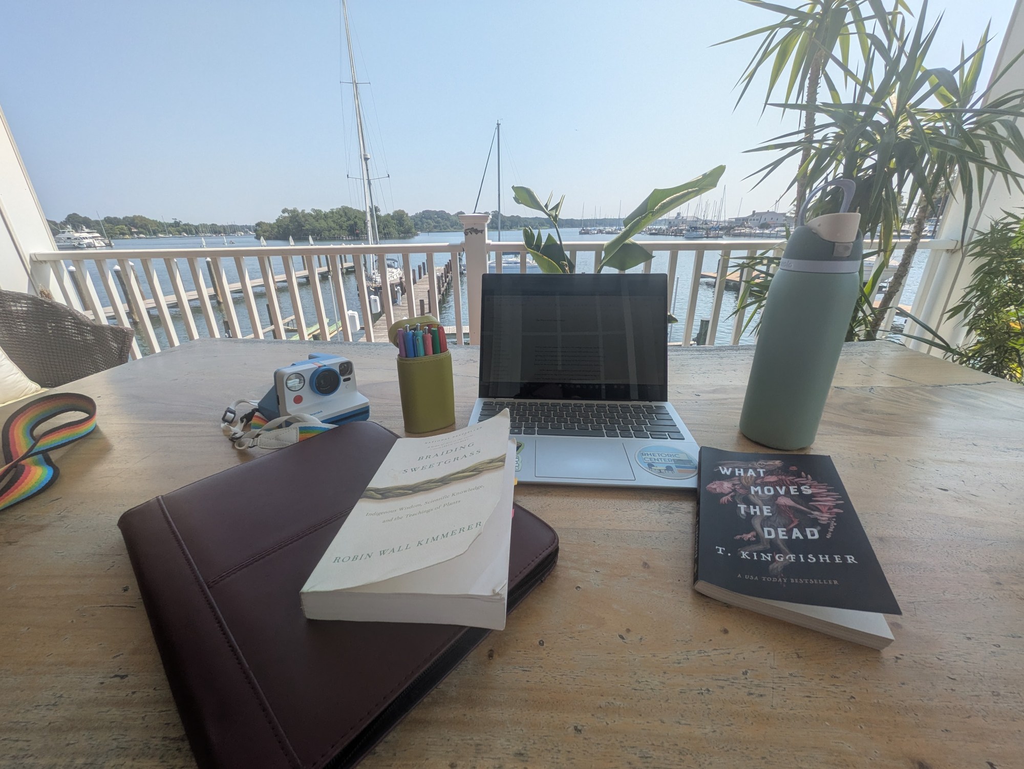 A laptop, some books, a polaroid sit on a table on a porch overlooking the water.